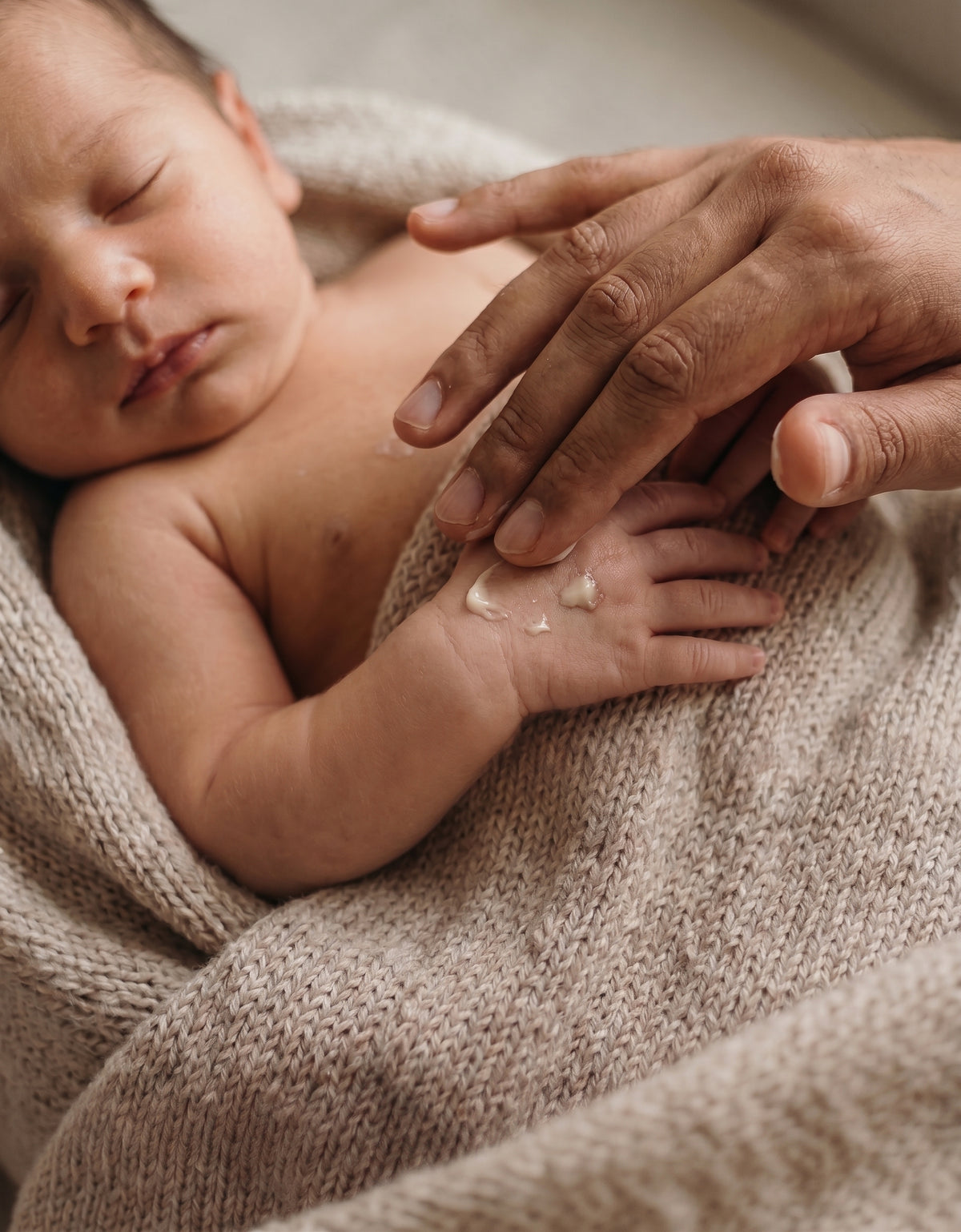 A close-up lifestyle shot of a sleeping newborn baby wrapped in a beige knit blanket, with an adult hand gently touching the baby's hand where a small dollop of white cream is visible.