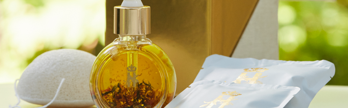 A round glass dropper bottle filled with golden yellow Balancing Rose facial serum by Royal Heir Botanicals, shown alongside white sachets with gold lettering with botanical soaks and a white konjac sponge on a light surface, with a blurred green and gold background.