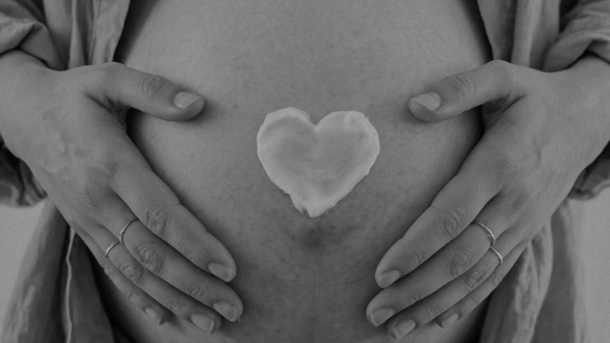 Black and white close-up of a pregnant woman's bare belly with both hands resting on the sides and a heart-shaped body butter centered on the abdomen.