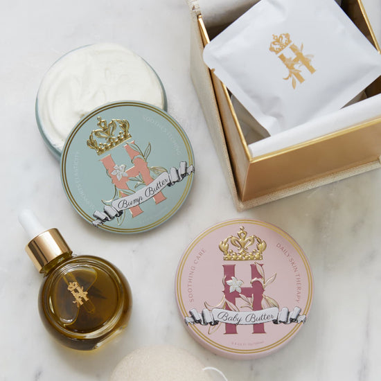 A luxury maternity skincare set arranged on a white marble surface. The set includes a small amber glass dropper bottle with Tender skin facial serum featuring a gold logo, two round decorative tin containers with whipped organic body butters, a beige box for white botanical soak sachets  with gold logo laying on top of it, and a white konjac sponge.