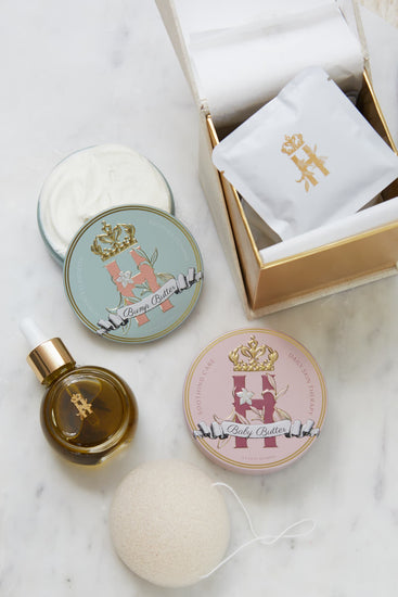 A luxury maternity skincare set arranged on a white marble surface. The set includes a small amber glass dropper bottle with Tender skin facial serum featuring a gold logo, two round decorative tin containers with whipped organic body butters, a beige box for white botanical soak sachets  with gold logo laying on top of it, and a white konjac sponge.