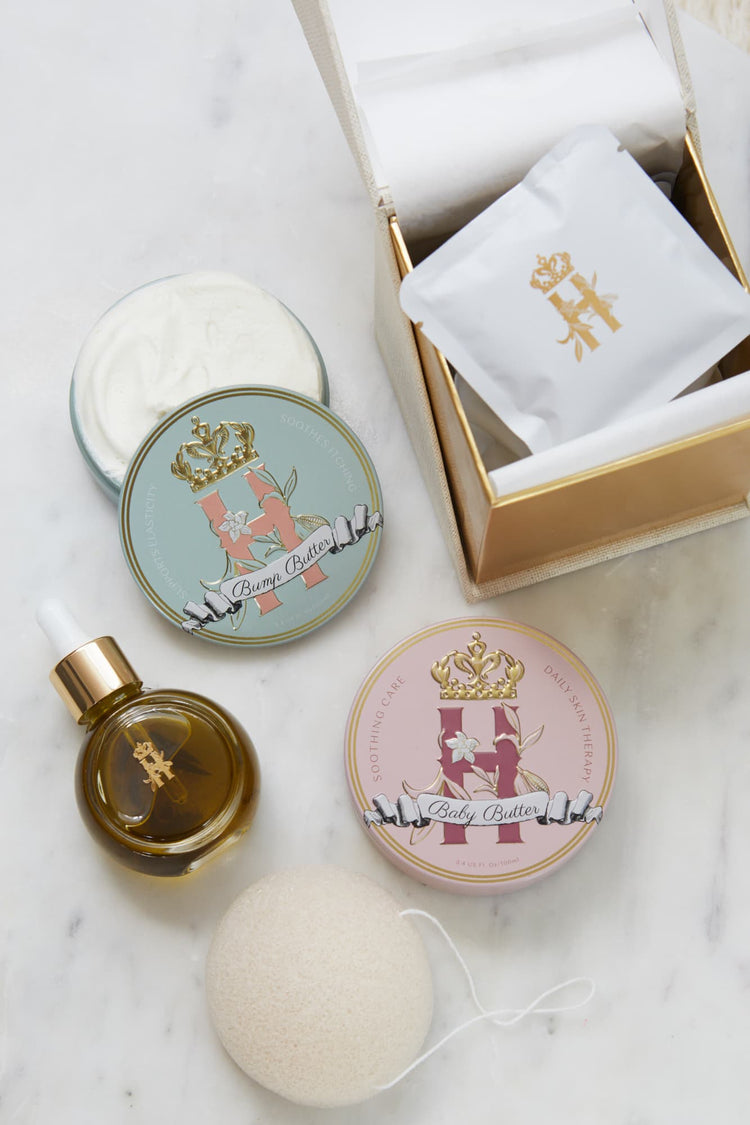 A luxury maternity skincare set arranged on a white marble surface. The set includes a small amber glass dropper bottle with Tender skin facial serum featuring a gold logo, two round decorative tin containers with whipped organic body butters, a beige box for white botanical soak sachets  with gold logo laying on top of it, and a white konjac sponge.