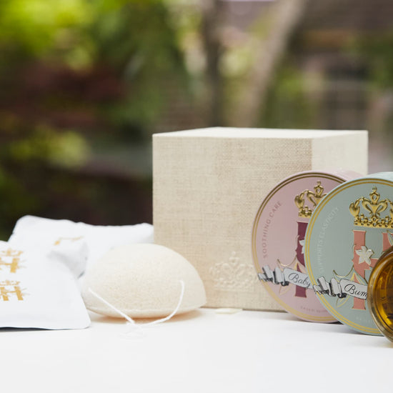 A luxury maternity skincare set arranged on a white surface with a blurred green outdoor background. The set includes a small amber glass dropper bottle with Tender skin facial serum, two round decorative tin containers with whipped organic body butters, a beige box for white botanical soak sachets  with gold logo laying next to it, and a white konjac sponge.