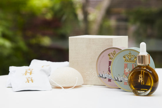 A luxury maternity skincare set arranged on a white surface with a blurred green outdoor background. The set includes a small amber glass dropper bottle with Tender skin facial serum, two round decorative tin containers with whipped organic body butters, a beige box for white botanical soak sachets  with gold logo laying next to it, and a white konjac sponge.