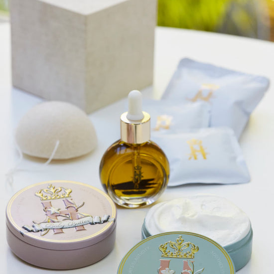 A luxury maternity skincare set arranged on a white surface with a blurred green outdoor background. The set includes a small amber glass dropper bottle with Tender skin facial serum, two round decorative tin containers with whipped organic body butters, a beige box for white botanical soak sachets  with gold logo laying next to it, and a white konjac sponge.