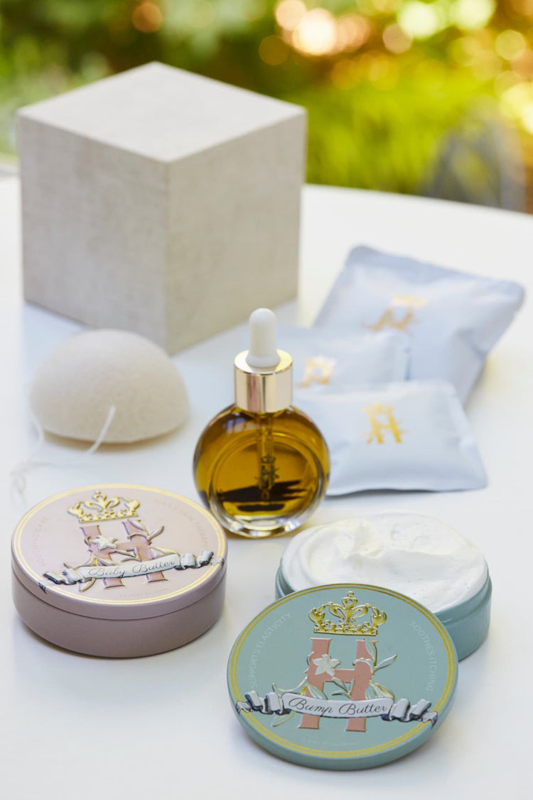 A luxury maternity skincare set arranged on a white surface with a blurred green outdoor background. The set includes a small amber glass dropper bottle with Tender skin facial serum, two round decorative tin containers with whipped organic body butters, a beige box for white botanical soak sachets  with gold logo laying next to it, and a white konjac sponge.