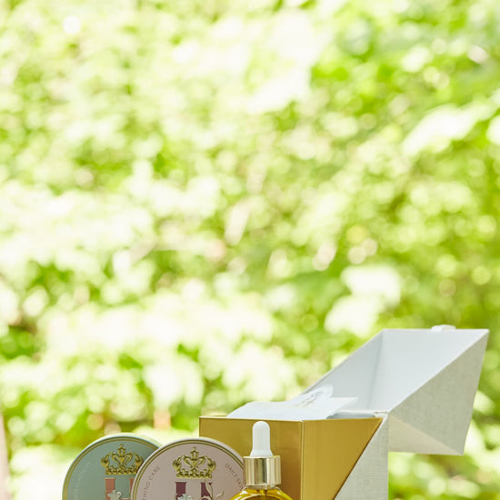 A luxury maternity skincare set arranged on a white surface with a blurred green outdoor background. The set includes a small amber glass dropper bottle with Tender skin facial serum, two round decorative tin containers with whipped organic body butters, a beige box for white botanical soak sachets  with gold logo laying next to it, and a white konjac sponge.