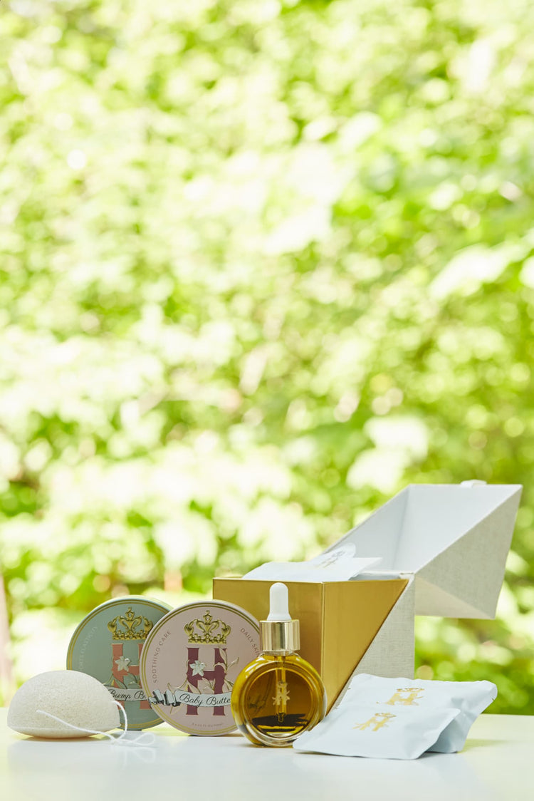A luxury maternity skincare set arranged on a white surface with a blurred green outdoor background. The set includes a small amber glass dropper bottle with Tender skin facial serum, two round decorative tin containers with whipped organic body butters, a beige box for white botanical soak sachets  with gold logo laying next to it, and a white konjac sponge.