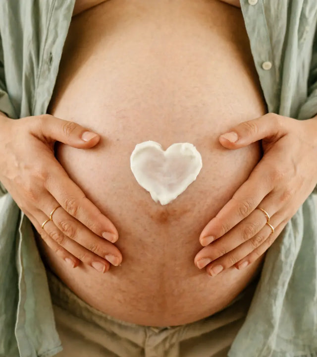 A close-up of a pregnant woman's bare belly with both hands resting on it, wearing an open sage green shirt. A small white heart-shaped cosmetic butter is placed on the center of the belly.