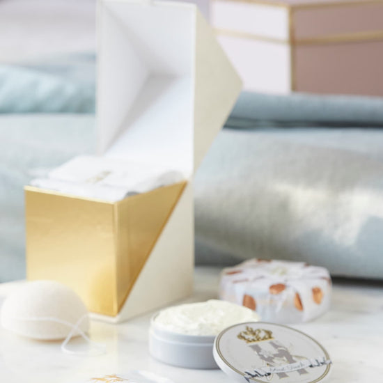 Luxury skincare set on white marble surface with blurred pastel pink and blue  background featuring a cream and gold cube shaped box open with a konjac sponge with a string, round tin with a crest logo and a gold crown holding ROyal Heir Botanicals whipped organic, EWG verified First Touch Butter for very delicate skin and a couple of white sachets with lavender botanical soak and a gold Royal Heir logo.