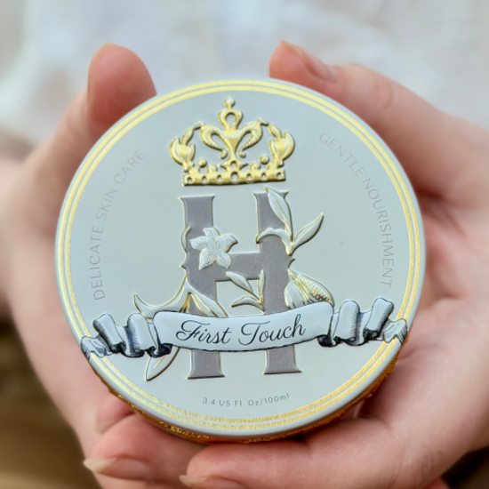 Blue cosmetic tin by Royal Heir Botanicals with a grey H logo with a golden crown above it, First Touch butter, EWG verified, organic formula for newborns held by woman's hands displaying the front to the camera