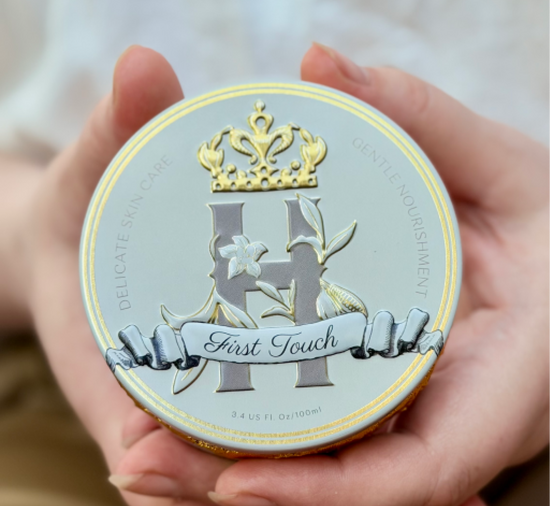 Blue cosmetic tin by Royal Heir Botanicals with a grey H logo with a golden crown above it, First Touch butter, EWG verified, organic formula for newborns held by woman's hands displaying the front to the camera
