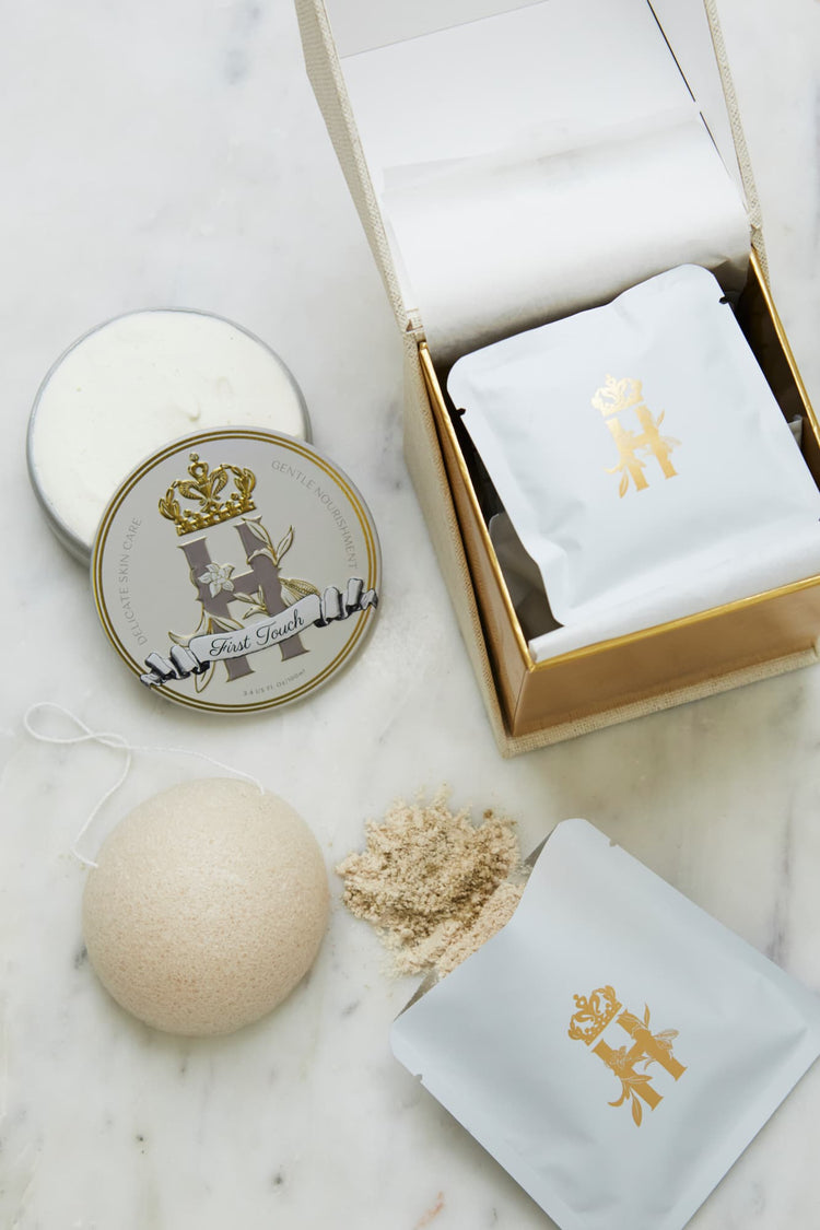 Luxury skincare set on white marble surface  featuring a cream and gold cube shaped box open with a konjac sponge with a string, round tin with a crest logo and a gold crown holding ROyal Heir Botanicals whipped organic, EWG verified First Touch Butter for very delicate skin and a couple of white sachets with lavender botanical soak and a gold Royal Heir logo.