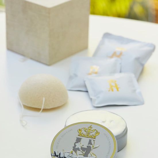 Luxury skincare set on white surface with blurred green foliage background featuring a cream and gold cube shaped box with a konjac sponge with a string, round tin with a crest logo and a gold crown holding ROyal Heir Botanicals whipped organic, EWG verified First Touch Butter for very delicate skin and a couple of white sachets with lavender botanical soak and a gold Royal Heir logo.