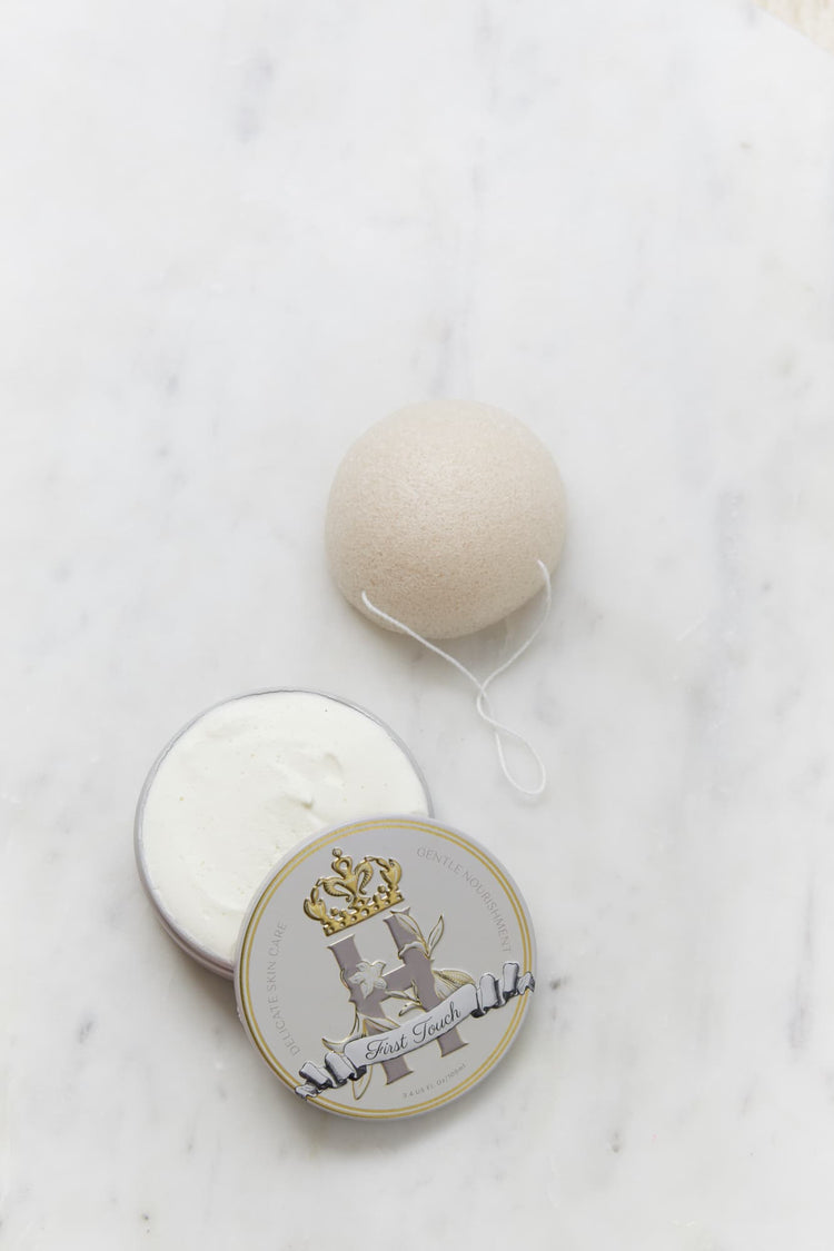 A round, off-white Konjac Sponge resting on a white marble surface next to its open warm grey tin, Royal Heir Botanicals First Touch butter, EWG verified, which features a decorative gold and grey crest label.