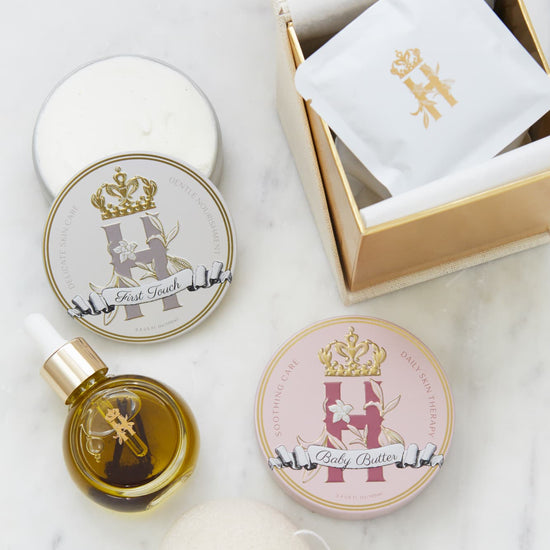 A luxury skincare gift set for a new mother and a baby by Royl Heir Botanicals arranged on a white marble surface, featuring a gold-toned box with a white botanical soak sachet, a round tin labeled 'Baby Butter in Pink', and a tin with First touch butter,  a small dropper bottle with facial serum, konjac exfoliating sponge.