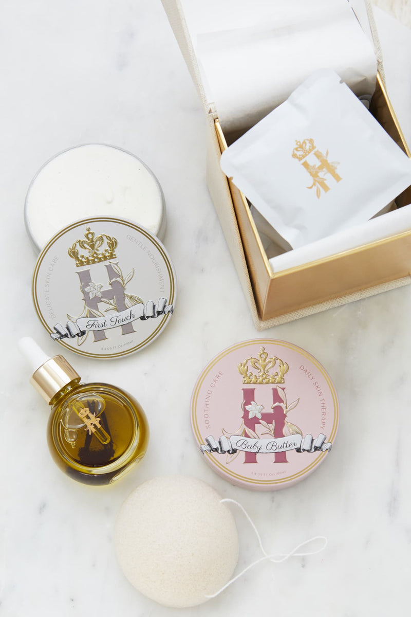 A luxury skincare gift set for a new mother and a baby by Royl Heir Botanicals arranged on a white marble surface, featuring a gold-toned box with a white botanical soak sachet, a round tin labeled 'Baby Butter in Pink', and a tin with First touch butter,  a small dropper bottle with facial serum, konjac exfoliating sponge.