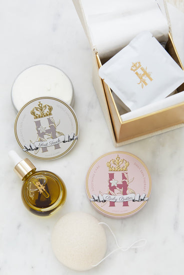 A luxury skincare gift set for a new mother and a baby by Royl Heir Botanicals arranged on a white marble surface, featuring a gold-toned box with a white botanical soak sachet, a round tin labeled 'Baby Butter in Pink', and a tin with First touch butter,  a small dropper bottle with facial serum, konjac exfoliating sponge.