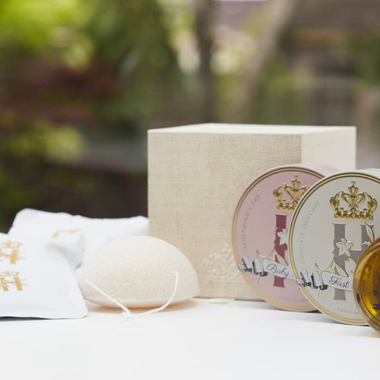 A skincare set featuring a round glass dropper bottle filled with golden serum, two round metal tins with illustrated lids, a textured cream box, a white bath bomb, and two white pouches with gold characters, arranged on a white surface with a blurred green garden background.