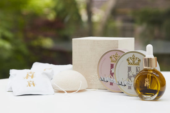 A skincare set featuring a round glass dropper bottle filled with golden serum, two round metal tins with illustrated lids, a textured cream box, a white bath bomb, and two white pouches with gold characters, arranged on a white surface with a blurred green garden background.