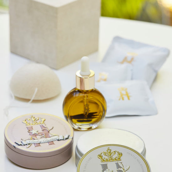A styled Royal Heir Botanicals aromatherapy neurosomatic skincare arrangement on a white surface featuring a dropper bottle with facial oil serum, two round metal tins with organic butter for mom and baby, white sachets with gold lettering holding botanical soak, and a beige box, photographed against a softly blurred outdoor green background.