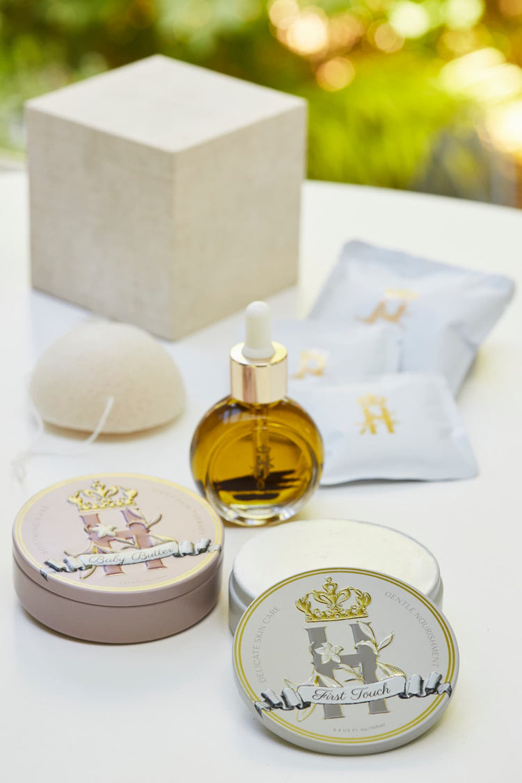 A styled Royal Heir Botanicals aromatherapy neurosomatic skincare arrangement on a white surface featuring a dropper bottle with facial oil serum, two round metal tins with organic butter for mom and baby, white sachets with gold lettering holding botanical soak, and a beige box, photographed against a softly blurred outdoor green background.