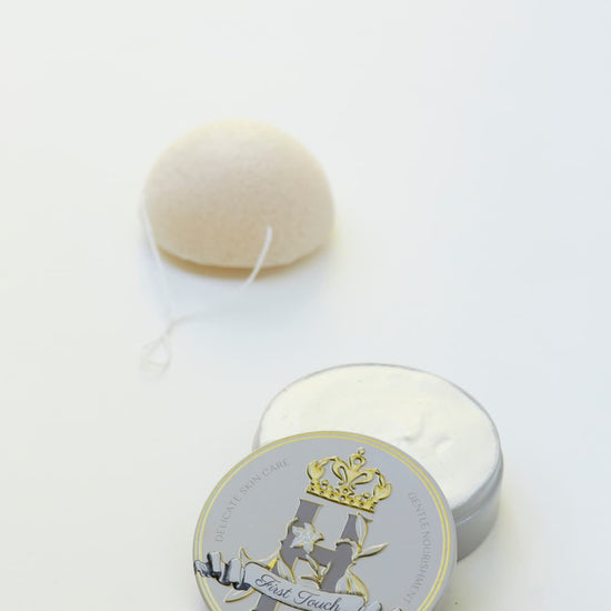 A small round metal tin with a warm gray lid featuring a gold crown and the words 'First Touch' sits beside an open tin of whipped butter, with a white konjac sponge in the background on a white surface.