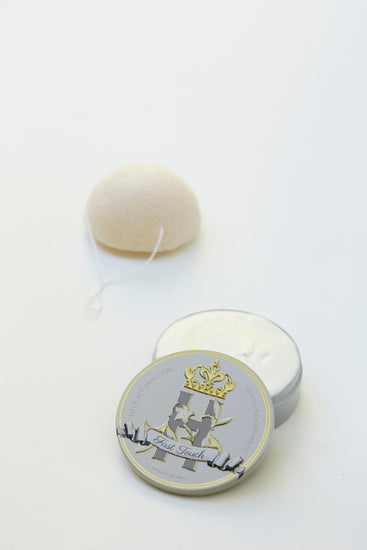 A small round metal tin with a warm gray lid featuring a gold crown and the words 'First Touch' sits beside an open tin of whipped butter, with a white konjac sponge in the background on a white surface.