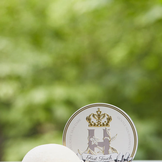 A round white Konjac sponge with a white string lies beside a round white tin labeled First Touch, set against a soft, blurred green outdoor background.