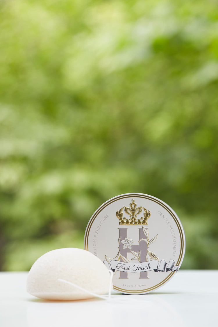 A round white Konjac sponge with a white string lies beside a round white tin labeled First Touch, set against a soft, blurred green outdoor background.
