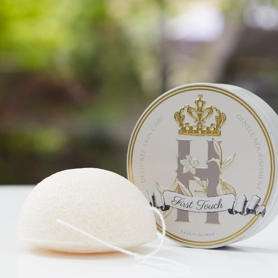A round white Konjac  sponge with a white string loop sits in front of a round white and gold branded tin with ROyal Heir Botanicals First Touch butter, EWG verified on a white surface with a blurred green outdoor background. 
