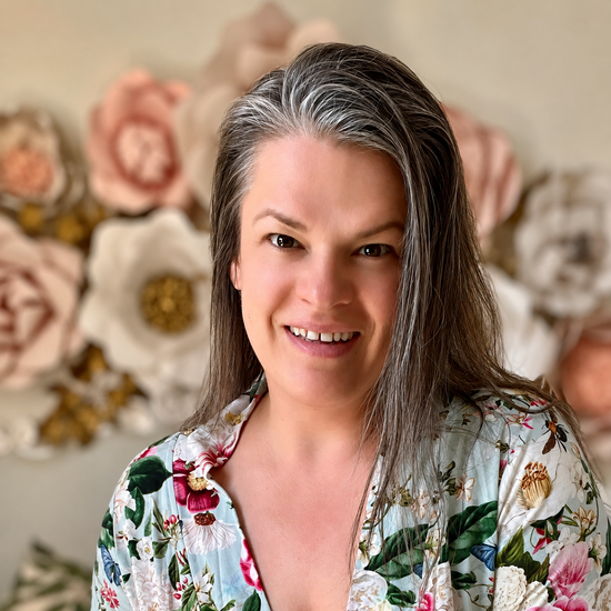 Portrait of Henrieta, former nurse, certified clinical aromatherapist and a somatic experiencing practitioner photographed against a wall with blush and white colored paper flowers.