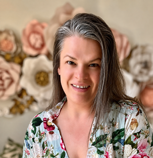 Portrait of Henrieta, former nurse, certified clinical aromatherapist and a somatic experiencing practitioner photographed against a wall with blush and white colored paper flowers.