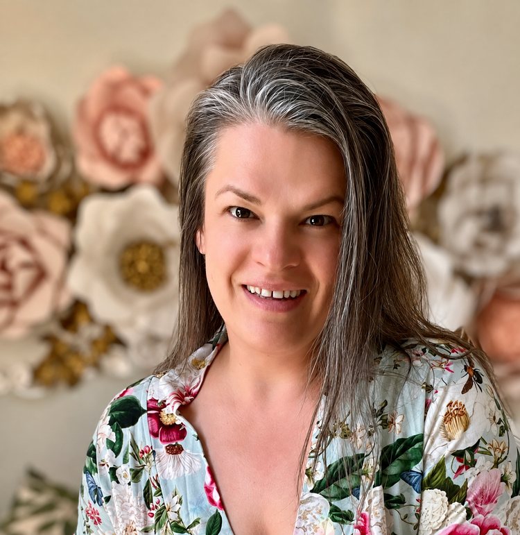 Portrait of Henrieta, former nurse, certified clinical aromatherapist and a somatic experiencing practitioner photographed against a wall with blush and white colored paper flowers.