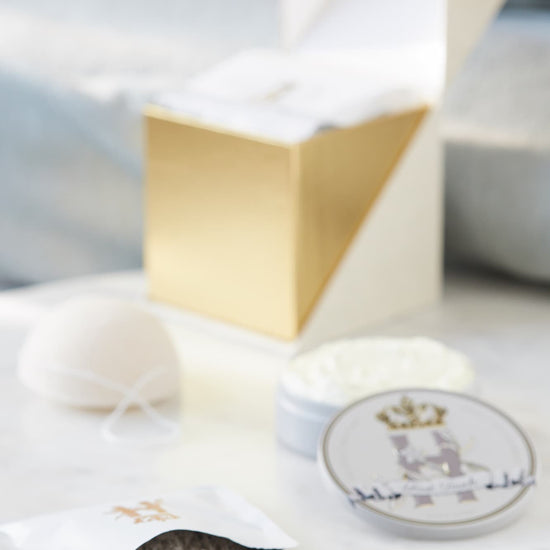 A luxury bath and body set featuring a white and gold geometric box, a white konjac sponge, a jar of white cream, a small round tin with Royal Heir Botanicals EWG verified First Touch butter with a crest, and a sachet with lavender botanical soak spilled on a white marble surface.