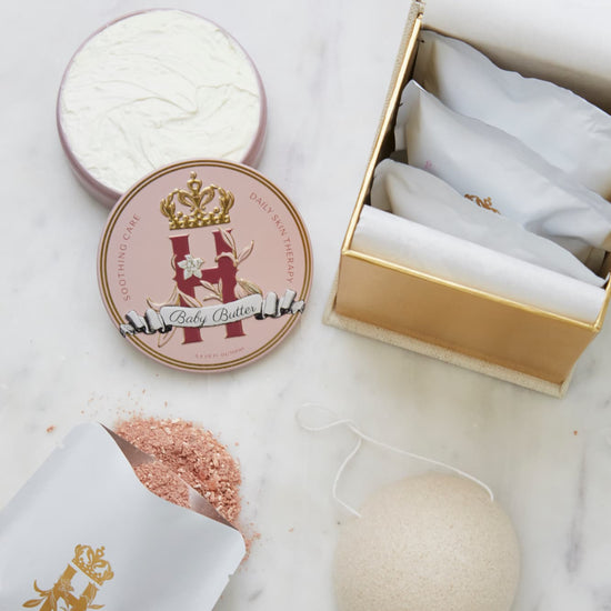 A luxury maternity skincare set arranged on a white marble surface. The set includes pink, round decorative tin container with whipped organic body butter, a beige box for white botanical soak sachets  with gold logo inside the box and laying next to it with rose botanical soak poured on the table surface and a white konjac sponge.