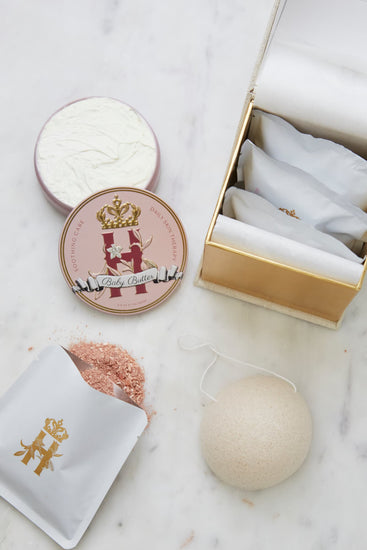 A luxury maternity skincare set arranged on a white marble surface. The set includes pink, round decorative tin container with whipped organic body butter, a beige box for white botanical soak sachets  with gold logo inside the box and laying next to it with rose botanical soak poured on the table surface and a white konjac sponge.