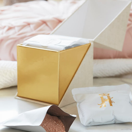 A luxury maternity skincare with a beige with gold interior box for white botanical soak sachets  with gold logo inside the box and laying next to it with rose botanical soak poured on the table surface with soft pink background and framed baby photos blurred. 