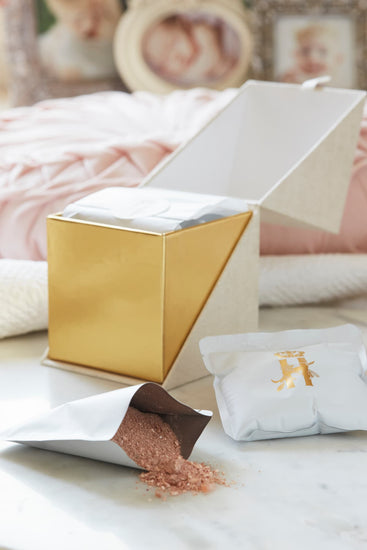 A luxury maternity skincare with a beige with gold interior box for white botanical soak sachets  with gold logo inside the box and laying next to it with rose botanical soak poured on the table surface with soft pink background and framed baby photos blurred. 