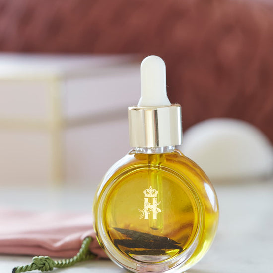 A round glass dropper bottle filled with golden yellow Tender skin facial serum by Royal Heir Botanicals, featuring a gold cap and a white rubber pipette. The bottle has a gold monogram logo on the front and is set on a white marble surface with a pink fabric and a blurred brown background.
