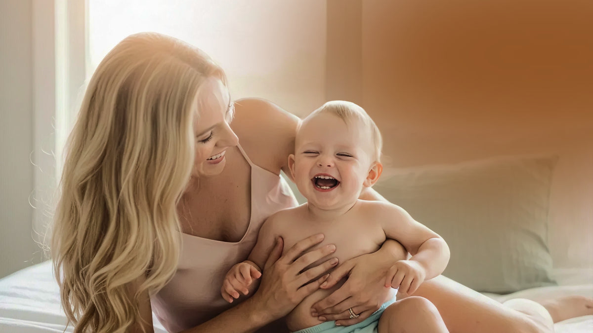 A smiling blonde mother holding and laughing with a baby in a softly lit bedroom setting.
