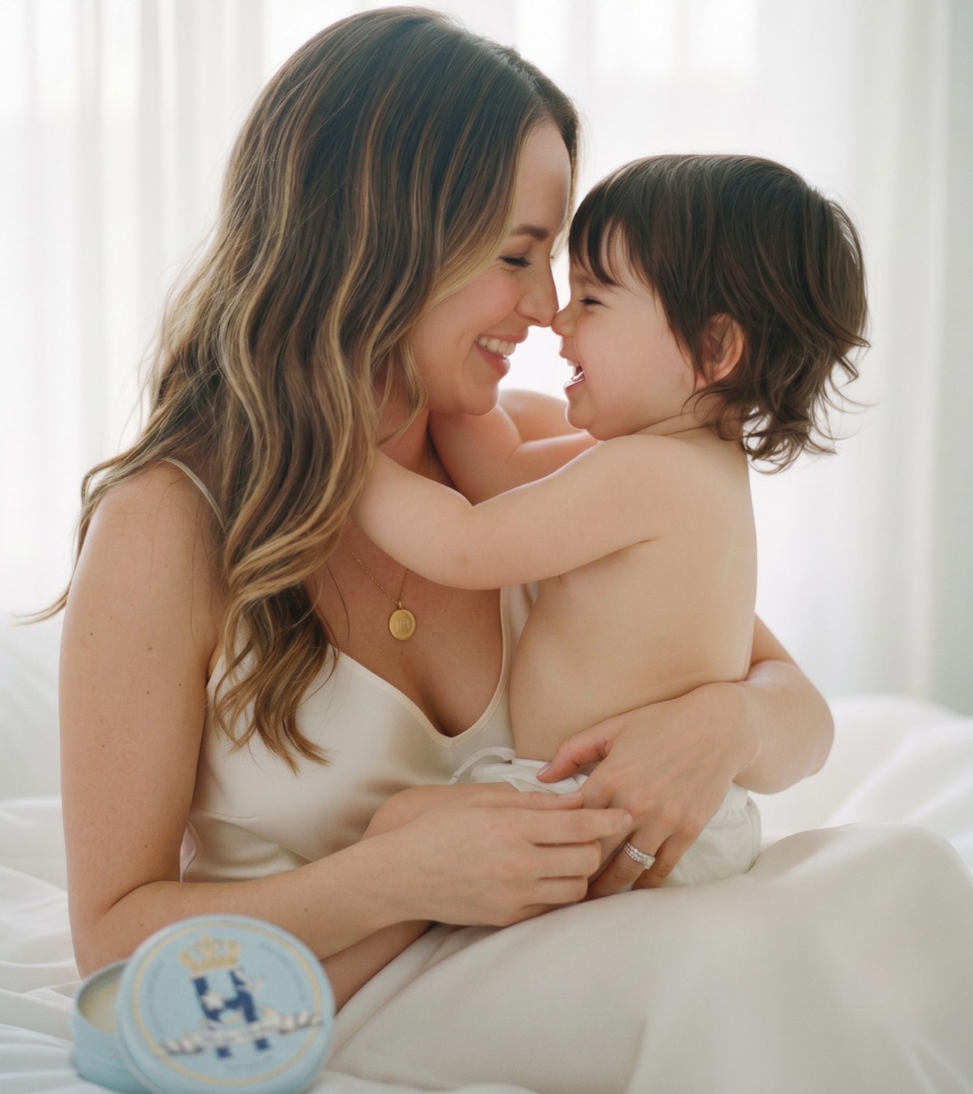 A woman in a cream satin slip dress holds a smiling toddler in a bright bedroom setting. A round light blue baby keepsake box with a gold crown and monogram is visible in the lower left corner.