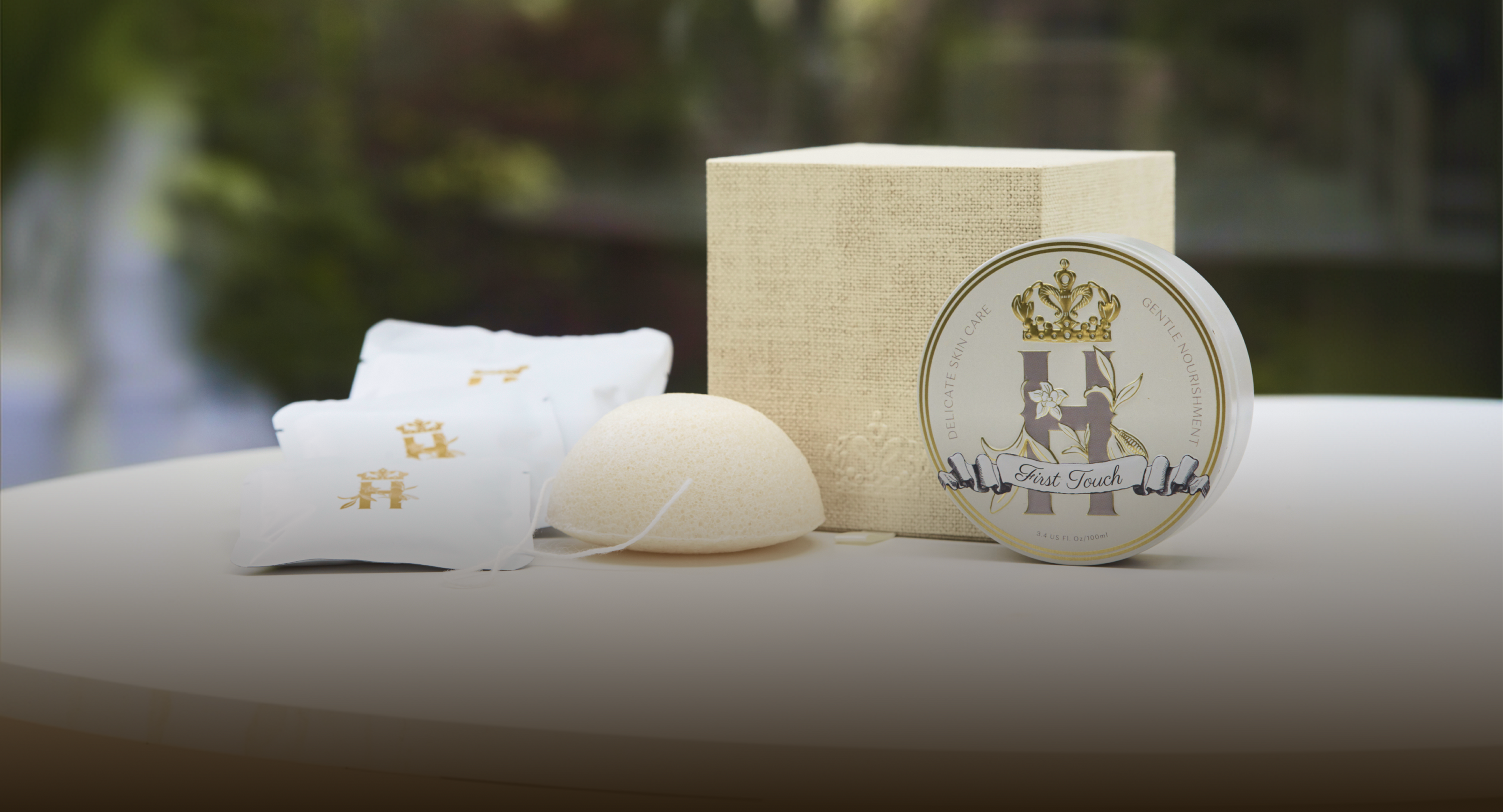 Evening containment ritual by Royal Heir Botanicals is a luxury maternity skincare set for mom and baby featuring a cream coloured cube shaped box for Lavender botanical soak, a round grey and gold tin with First touch whipped organic,EWG verified butter for newborns, konjac sponge with a string and a few white sachets with golden logo on the front with lavender botanical soak photographed on a white surface with blurred garden in the backgroun