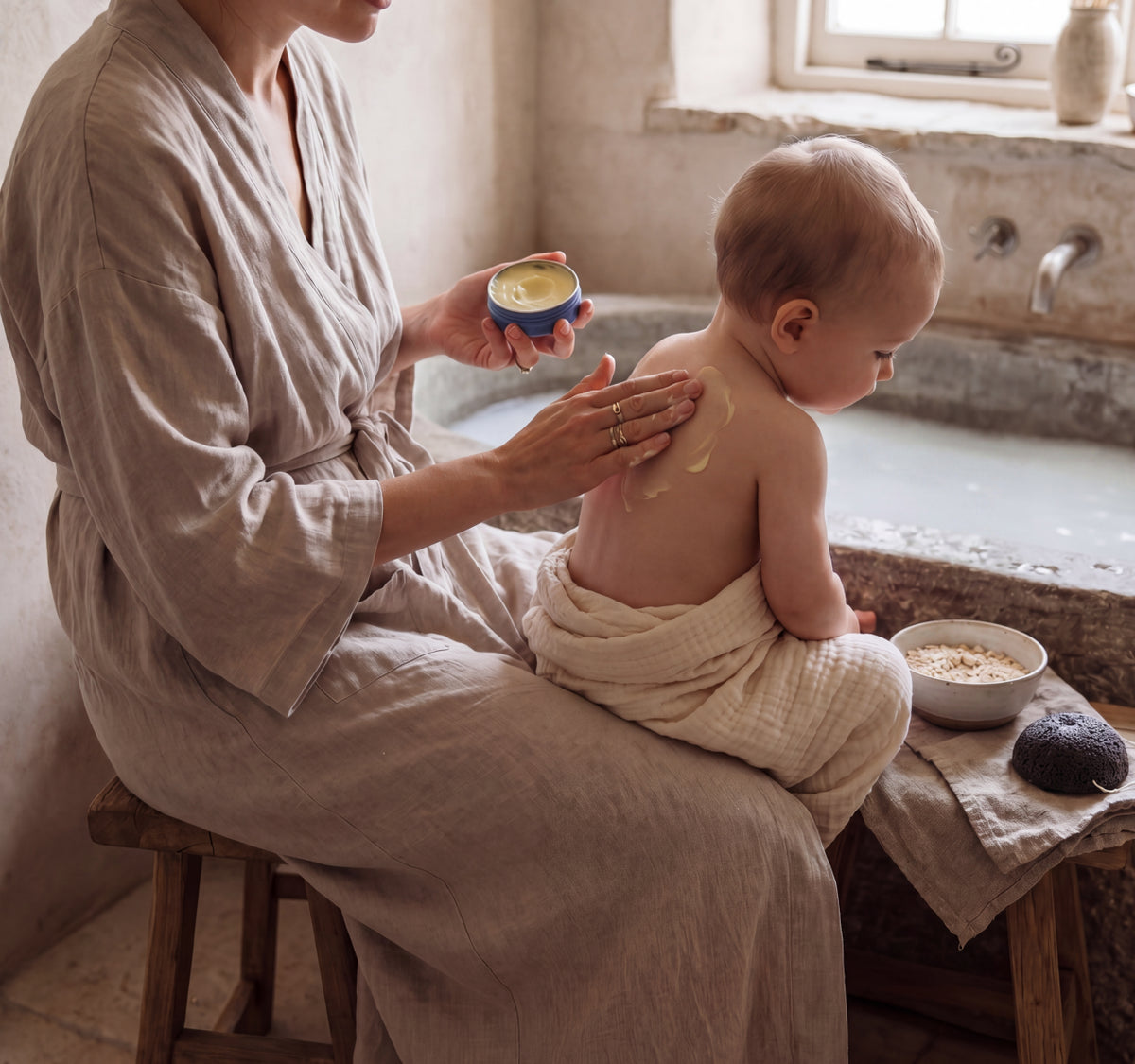 Mother massaging Royal Heir Botanicals Butter in Blue into her toddler's back as he sits on her lap in a bathroom with a filled up bathtub