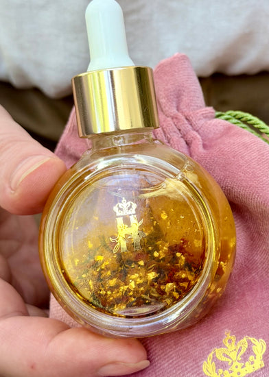 A hand holds a small round glass dropper bottle filled with golden amber Balancing  Rose Facial serum and embedded gold flakes, with a pink drawstring pouch and a little gold crown logo behind it
