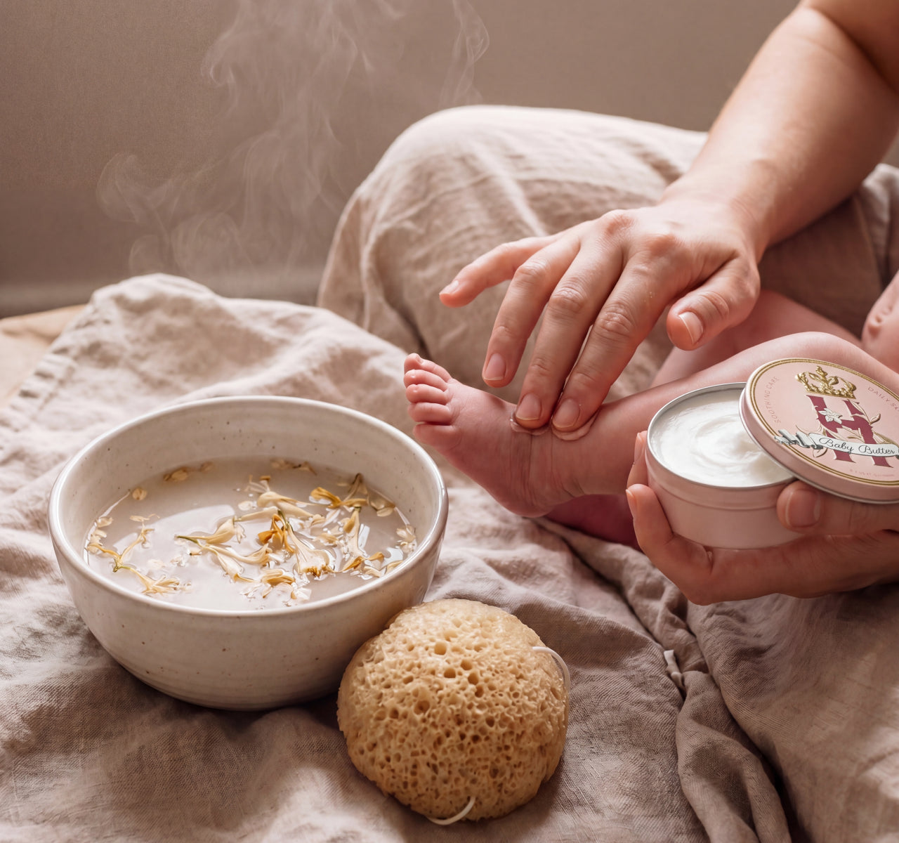 warm bathroom scene with a bowl with Honeysucle and Oat botanical soak, a sponge and a mother massaging whipped butter in pink by Royal Heir Botanicals into her baby's little foot.