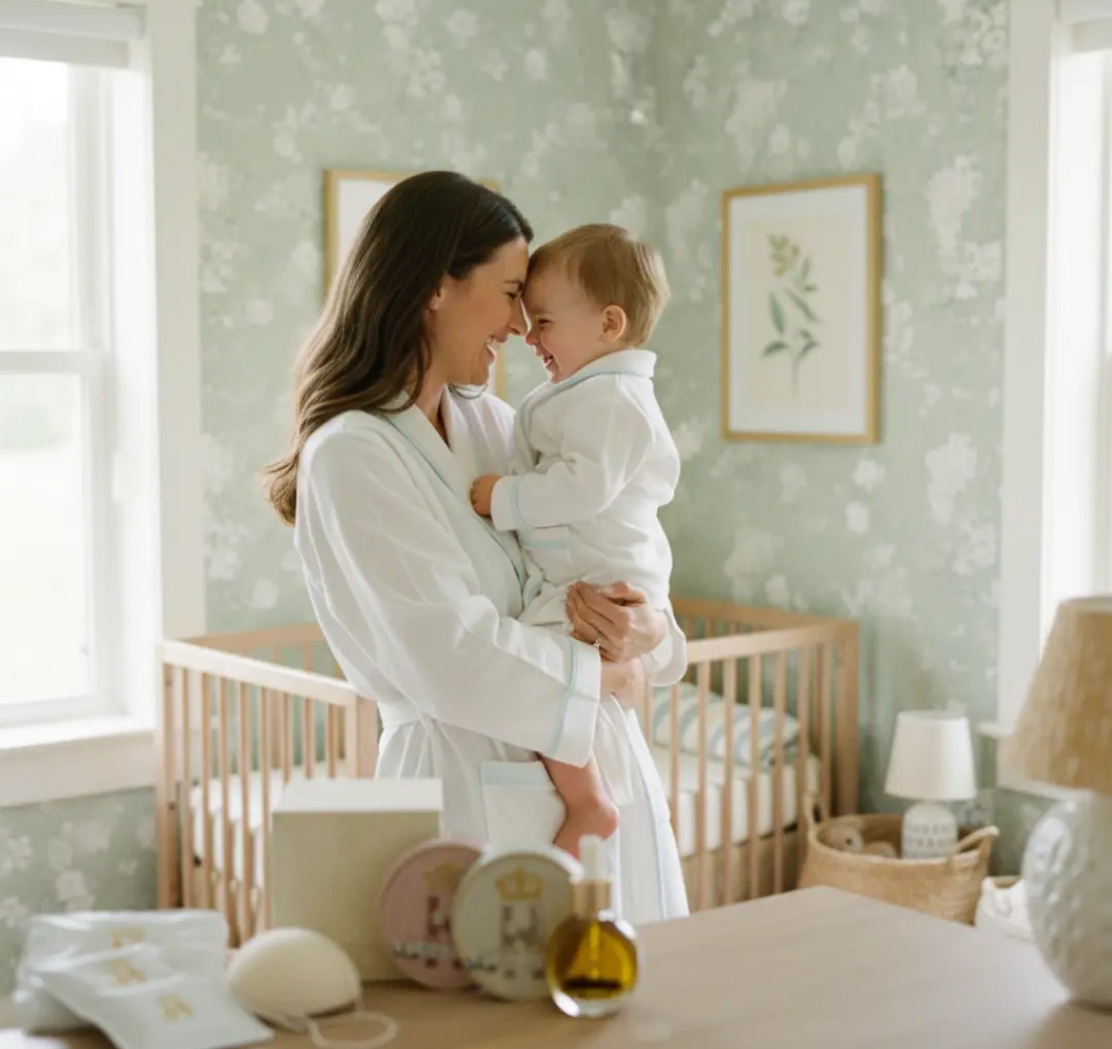 A mother in a white robe holds a baby dressed in a matching white outfit in a softly lit nursery with pale green floral wallpaper, a wooden crib, framed wall art, and bedside decor.