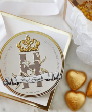 cosmetic round tin with Royal Heir Botanicals First touch butter on a white box eith golden edges and gold foil wrapped chocolate hearts next to it. 