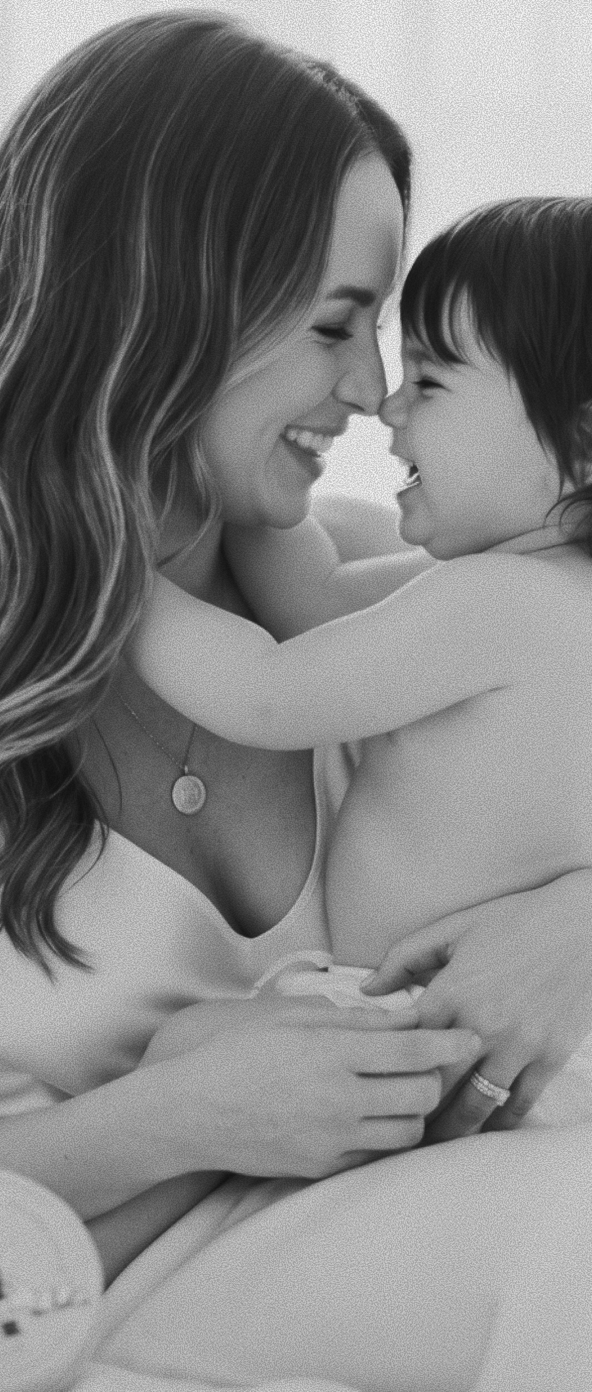 A black and white photograph of a smiling mother holding and embracing a baby, with the woman wearing a delicate round pendant necklace.