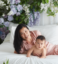 A smiling woman in a pink satin dress lies on a white bed with a baby beside her, with a large floral arrangement in the background.